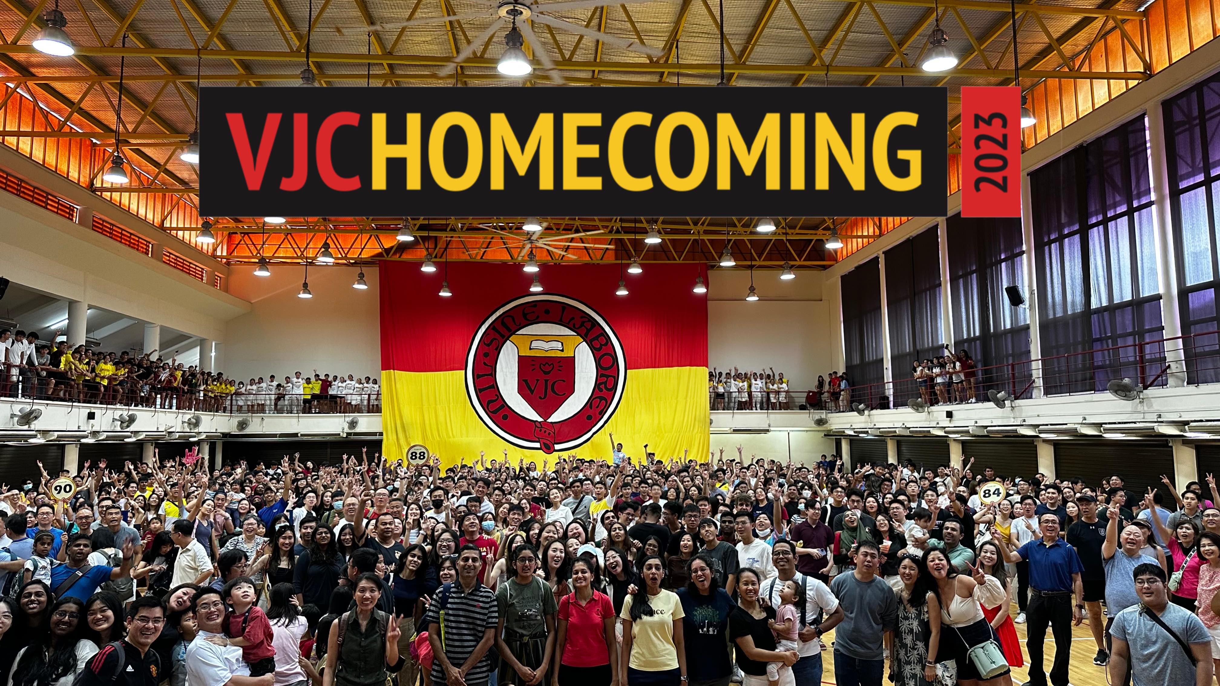 Large crowd gathered in a school hall for VJC Homecoming 2023, with a red and yellow VJC banner behind them.
